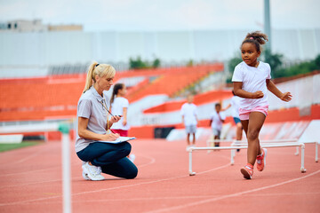 Obraz premium Sports teacher using stopwatch while black girl is jumping over hurdles on running track