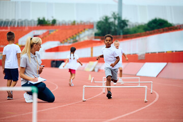 Black boy and his friends running on track with obstacles during exercise class at athletics club. - obrazy, fototapety, plakaty