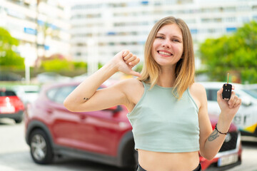 Young blonde woman holding car keys at outdoors proud and self-satisfied