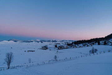 Fototapeta na wymiar Pink-blue sunset over a snow-covered village on the edge of a forest in a mountain valley