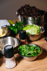 Fresh Greens and Salt Shakers on Kitchen Counter. Kitchen prep scene with bowls of fresh lettuce, cilantro, and other greens, accompanied by salt and pepper shakers on a wooden countertop.