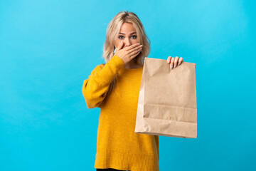 Young Russian woman holding a grocery shopping bag isolated on blue background covering mouth with...