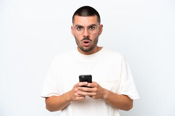 Young caucasian man isolated on white background looking at the camera while using the mobile with surprised expression