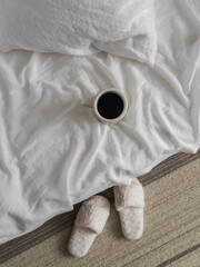 Coffee mug on the bed with white linen bedding, fur slippers on the carpet, top view