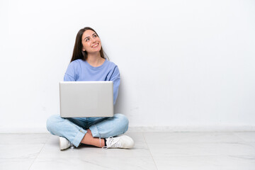 Young caucasian woman with laptop sitting on the floor isolated on white background looking up while smiling