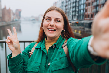 Happy 30s Women taking selfie on urban background. Young beautiful girl say Hi. Gdansk old town and famous Zuraw crane, A beautiful redhead woman is standing near Motlawa river, Traveler