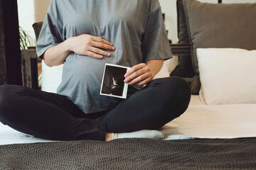 Cropped shot of pregnant woman holding pregnancy ultrasound or baby sonogram while sitting on bed