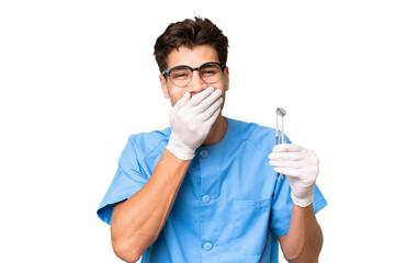 Young dentist man holding tools over isolated background happy and smiling covering mouth with hand