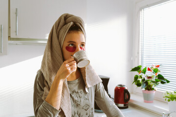 teenage girl, after shower, wearing patches under eyes, drinks coffee in home kitchen.