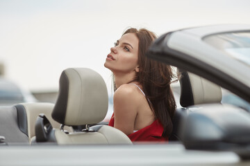 Young brunette sitting in convertible car and dreamily looking at sky