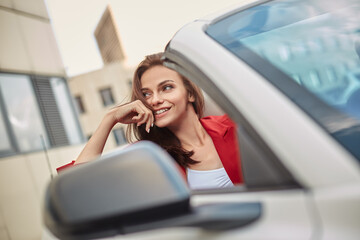 Cheerful playful young girl in red blazer enjoying summer day in convertible