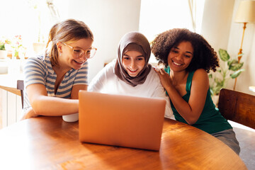 Three pretty female teenagers of different nationalities are looking at the laptop screen and laughing - obrazy, fototapety, plakaty