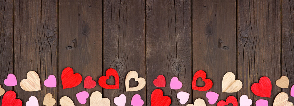 Valentines Day bottom border of wooden hearts. Top view on a dark wood banner background. Copy space.