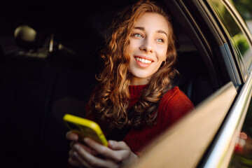 Positive young woman in casual clothes sits in the back seat of a car with a seat belt fastened using a mobile phone. Technology concept, traveling by car.