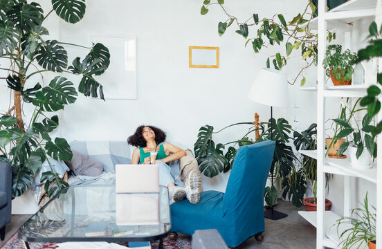 Charming ethnic african girl sits on sofa in front of laptop. Cute mixed race teenager put her feet on armchair and rests at home.