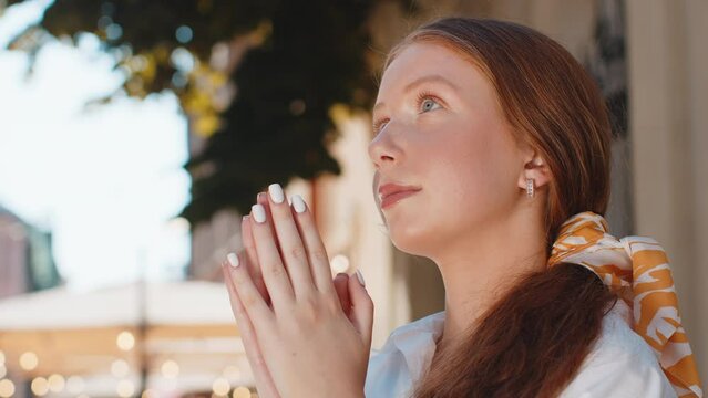 Portrait of red hair religion teenager girl praying closed eyes to God asking for blessing help forgiveness outdoors. Young woman clasping hands wishing luck in urban sunny city street. Town lifestyle