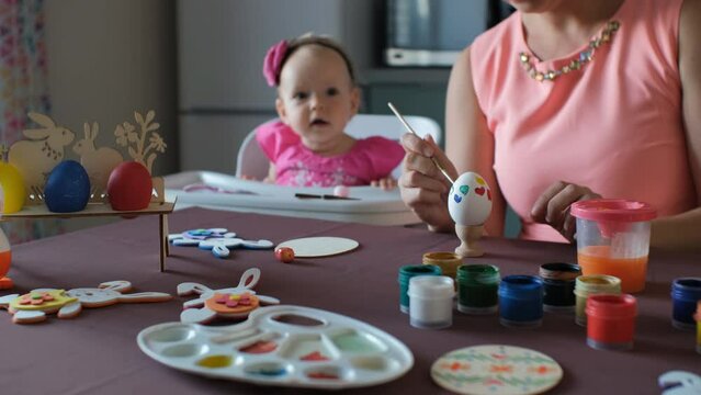 Cute baby girl watching her mother painting Easter egg in the kitchen, selective focus. Family preparing for Easter holidays