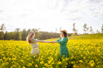 Naklejka na ściany i meble Two young women in beautiful dresses walk in a field with yellow blooming rapeseed. Beautiful girlfriends enjoying the weather, having fun in a flowering field. Concept of fun, relaxation.