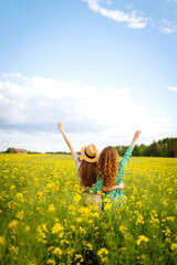 Two young women in beautiful dresses walk in a field with yellow blooming rapeseed. Beautiful...