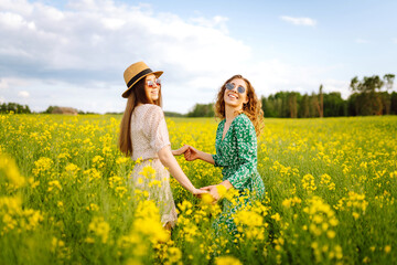 Fototapeta premium Two young women in beautiful dresses walk in a field with yellow blooming rapeseed. Beautiful girlfriends enjoying the weather, having fun in a flowering field. Concept of fun, relaxation.