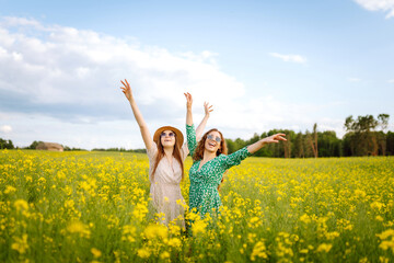 Naklejka na ściany i meble Two young women in beautiful dresses walk in a field with yellow blooming rapeseed. Beautiful girlfriends enjoying the weather, having fun in a flowering field. Concept of fun, relaxation.