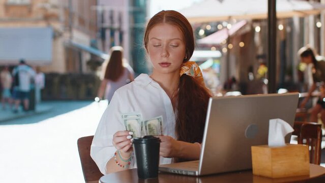 Happy rich freelancer girl counting money dollar cash, use calculator app, plans to order gifts food delivery online booking hotel room outdoors. Young woman tourist sitting at table in city cafeteria