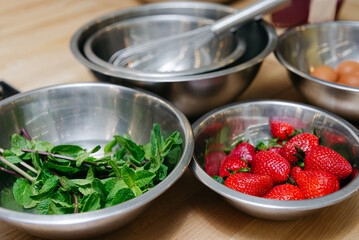 Fresh Strawberries and Mint in Stainless Steel Bowls. Ripe strawberries and vibrant green mint leaves in stainless steel bowls, ready for a culinary creation on a wooden kitchen surface.