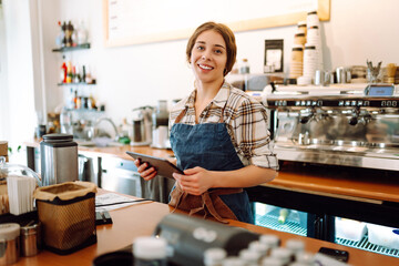 Portrait of a smiling cafe owner using a digital tablet behind the bar counter in a modern coffee shop. A young barista takes an order using a tablet. Small business concept. - obrazy, fototapety, plakaty