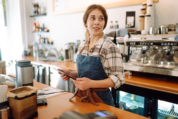 Portrait of a smiling cafe owner using a digital tablet behind the bar counter in a modern coffee shop. A young barista takes an order using a tablet. Small business concept. - Powered by Adobe