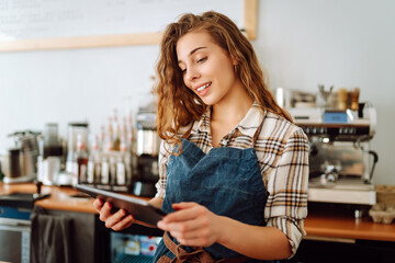 Portrait of a smiling cafe owner using a digital tablet behind the bar counter in a modern coffee shop. A young barista takes an order using a tablet. Small business concept.
