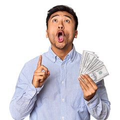 Young Chinese man holding dollar bills in studio pointing upside with opened mouth.