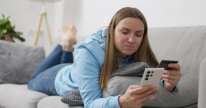 Woman relaxes on couch holding a smartphone and credit card, doing e-commerce
