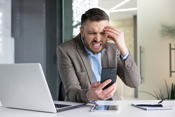Angry and disappointed young male businessman sitting in the office at the desk and looking at the phone screen.