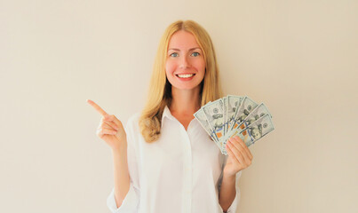 Portrait of happy smiling young woman holding cash money in dollar bills in her hands and points...