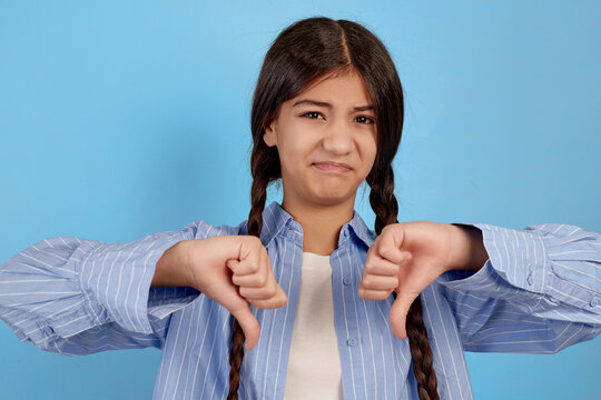 Malcontent teenage girl in blue shirt shows thumbs down on blue background.
