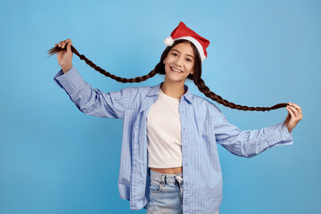 Smiling teenage girl with and Santa hat holding pigtails on blue background.