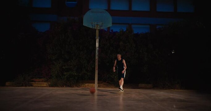 Basketball player run up to hoop and throw ball in, scoring a point. Nightime evening shot of player practice street basketball in outside court