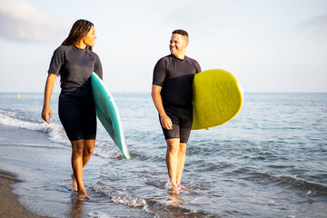 Two dark-skinned young men walk along the seashore dressed in wetsuits and carrying surfboards....