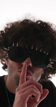 close up video of sexy cool man with spiked sunglasses in black hoodie making silence gesture and looking forward on grey background