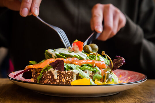 Salmon and avocado on bread with salad, capers, tomatoes and sauce on a plate being eaten with a knife and fork