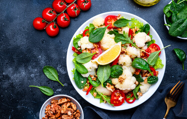 Healthy vegan salad with white cauliflower, baked red paprika, cherry tomatoes and spinach with walnuts, gray table background, top view