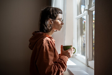Lonely sad teenager with cup of tea looking a window, brooding. Unsociable teen girl spending...