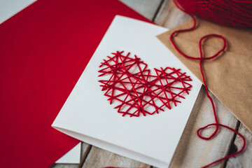 Concept of handmade simple holiday surprised for Saint Valentine's Day or Mother's Day. Easy to do DIY craft postcard from recycled paper with embroidering of red heart with wooden threads