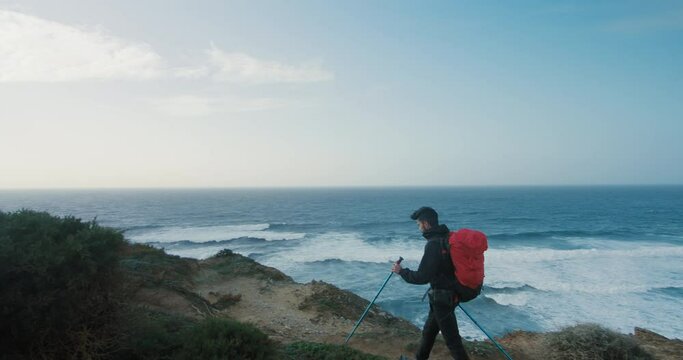 Side shot with gimball of hispanic young man walk on empty trail on cliffs along ocean. Windy hiking day in solitude. 