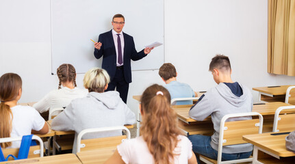 Male teacher standing at whiteboard in classroom, conducting lesson with teen students