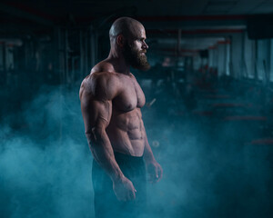 A muscular bald man poses shirtless in the dark amid smoke. A bodybuilder shows off his form in the...