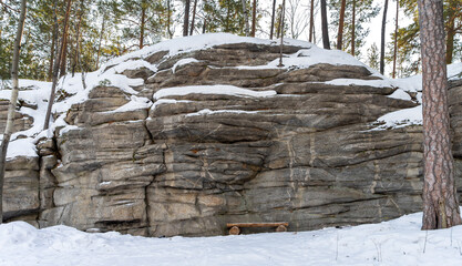 Wooden bench near rock Isetskaya wall. Granite array. Hiking in snow on winter day. Long rocky...