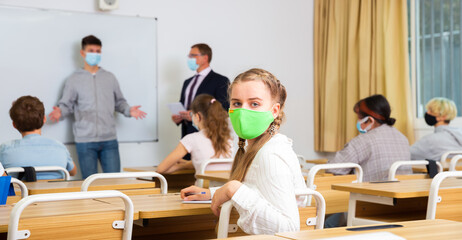 Portrait of diligent schoolgirl wearing protective face mask sitting in class working with...