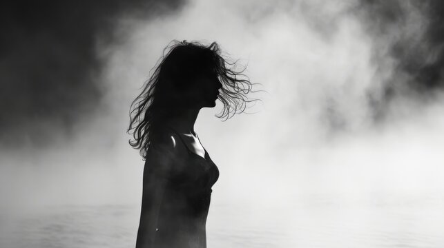  a black and white photo of a woman standing in a body of water with her hair blowing in the wind.