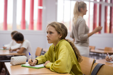 Focused school girl sitting at school desk in classroom on background with classmates